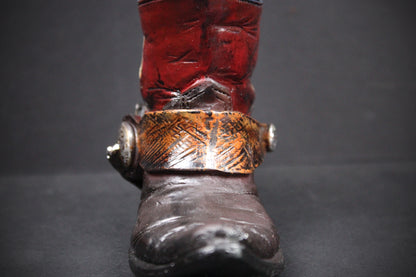 A close-up of a western-style boot with a dark brown leather foot, a wide metallic bronze-toned buckle strap, and a red leather shaft, set against a dark gray background.