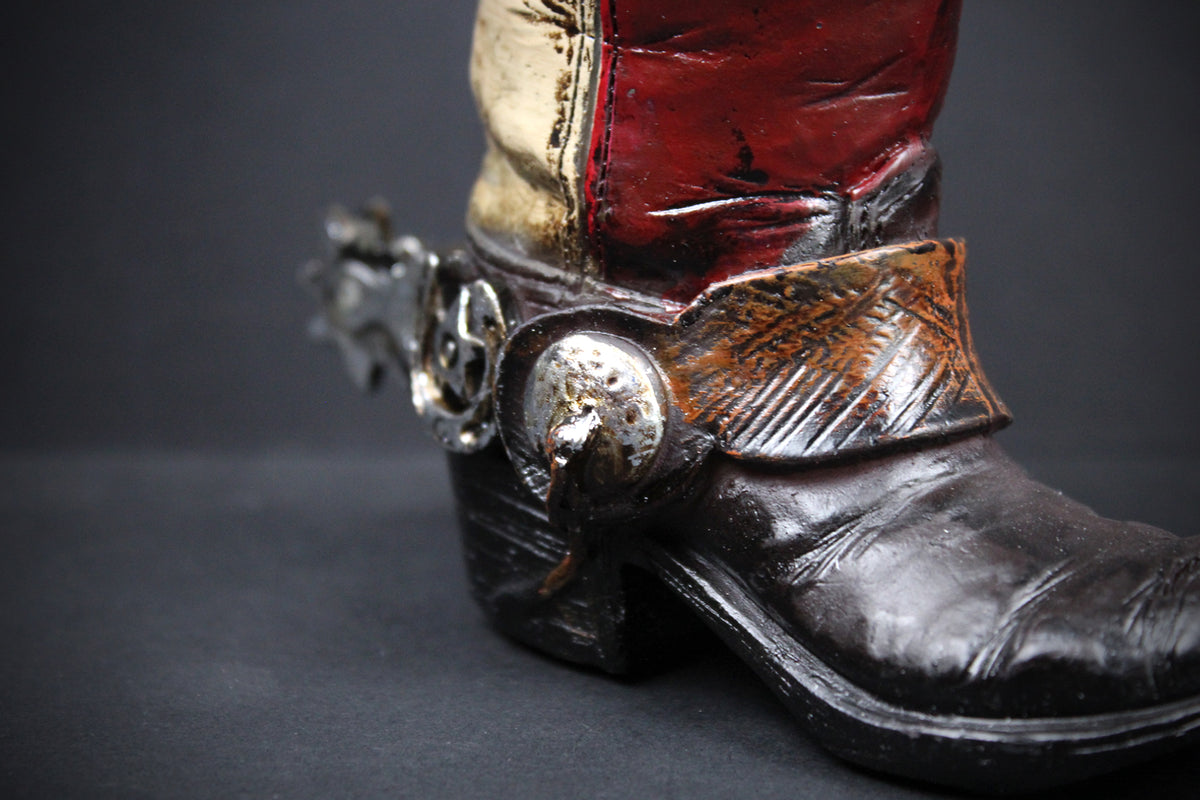 A close-up of a western-style boot with a dark brown leather foot, a red and beige shaft, and a decorative silver-toned spur and concho strap.