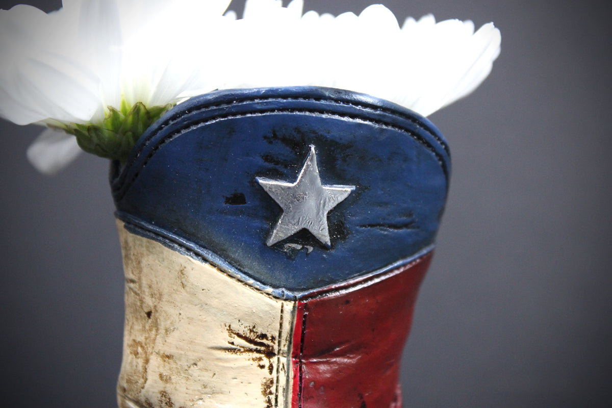 A close-up of a cowboy boot-shaped vase filled with white flowers, featuring a distressed blue, red, and white design with a silver star emblem on the front, set against a dark gray background.