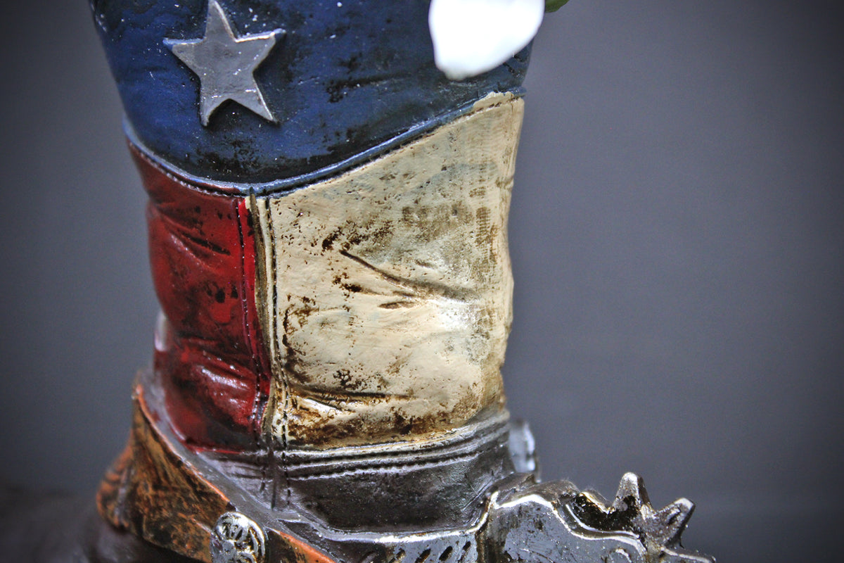 A close-up of a distressed cowboy boot figurine or sculpture, featuring a blue top section with a silver star, a red and off-white vertical stripe design, and a brown distressed leather lower section with a metal spur attached.