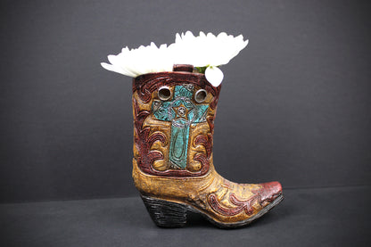 A miniature cowboy boot planter with a carved turquoise cross on the shaft, gold-toned detailing, and a white flower inserted into the opening, set against a dark gray background.