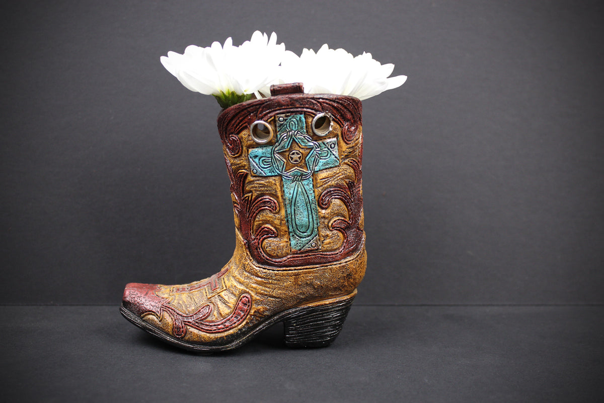 A miniature cowboy boot planter with a carved turquoise cross and floral details, filled with white daisies, set against a dark gray background.