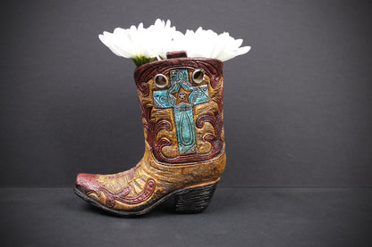 A miniature cowboy boot planter with a carved turquoise cross and floral details, filled with white daisies, set against a dark gray background.