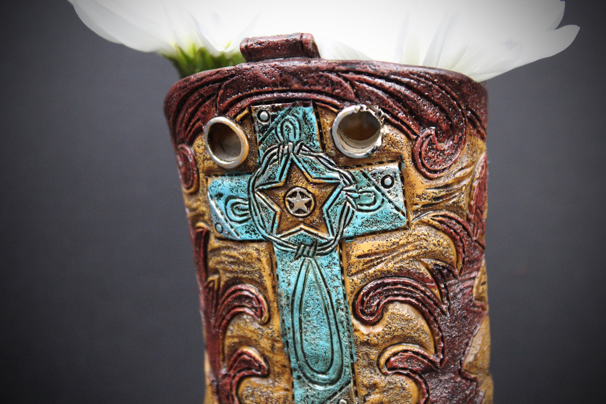 A close-up of a decorative ceramic pot or vase with a western-inspired design, featuring a turquoise and gold cross motif on a brown and gold embossed background, with a white flower stem emerging from the top against a dark gray background.