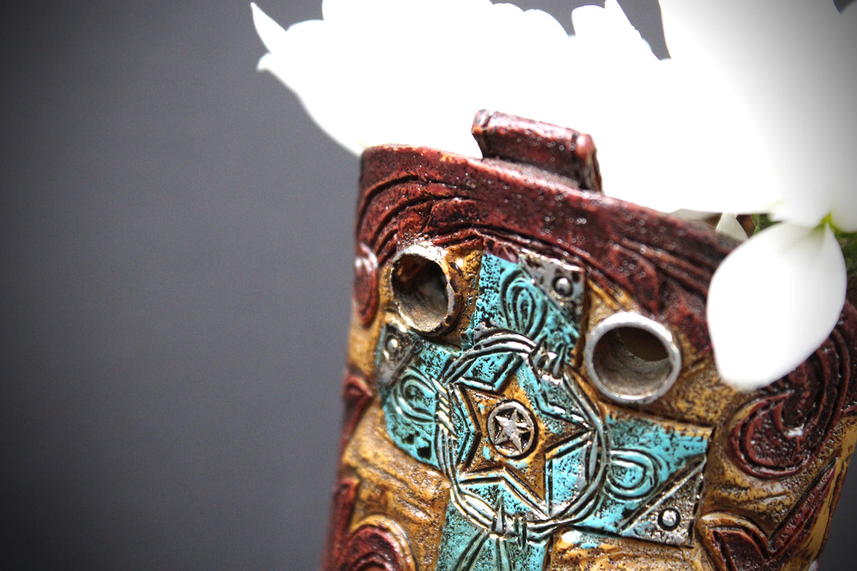 Close-up of a decorative ceramic planter with a western-inspired design, featuring turquoise, gold, and reddish-brown patterns, with white flowers emerging from the top against a dark gray background.