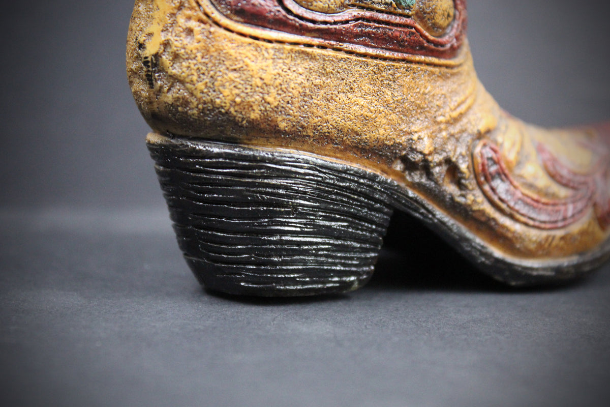 A close-up shot of a distressed western-style boot, showing the textured black heel and a weathered tan upper with red and gold decorative stitching, set against a dark gray background.