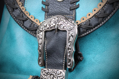 Close-up of a western-style leather belt with a large ornate silver buckle, black leather strap, tan braided trim, and a turquoise blue background.