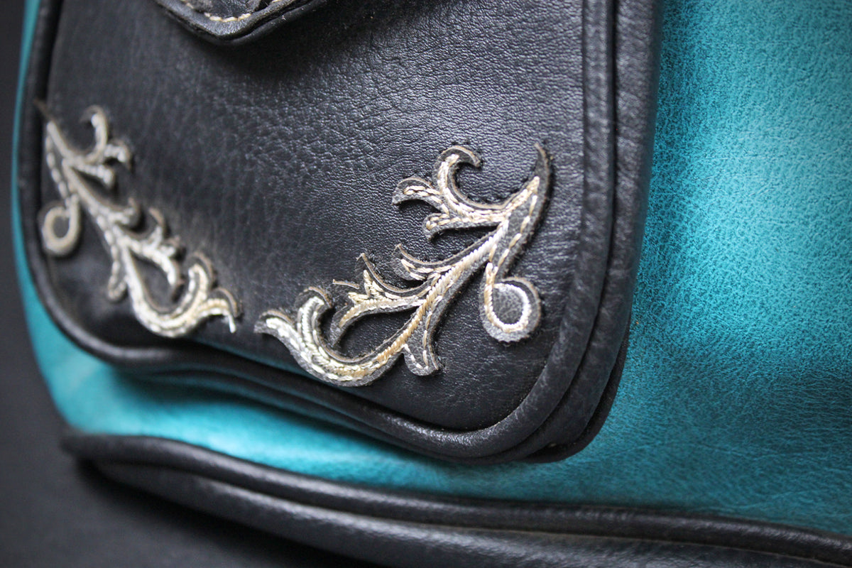 Close-up of a turquoise and black leather bag featuring ornate silver embroidered scrollwork on a black leather flap.