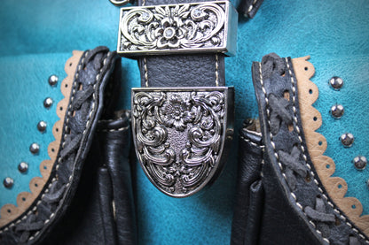 Close-up of a turquoise leather belt with a large ornate silver-toned buckle featuring floral scrollwork, flanked by black leather straps with tan scalloped trim and silver studs.