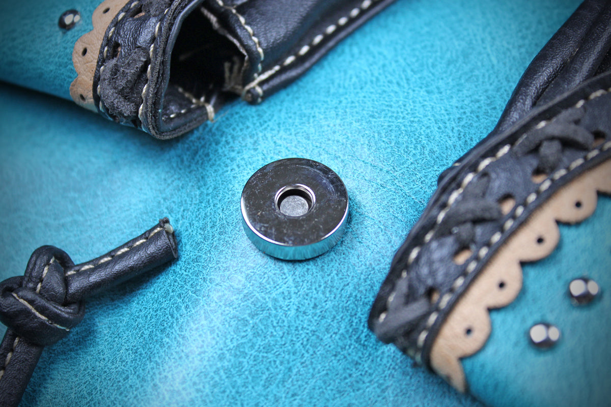 A close-up of a turquoise leather boot or shoe detail with black leather trim, white stitching, scalloped edges, and a central silver metal snap button on a textured turquoise surface.