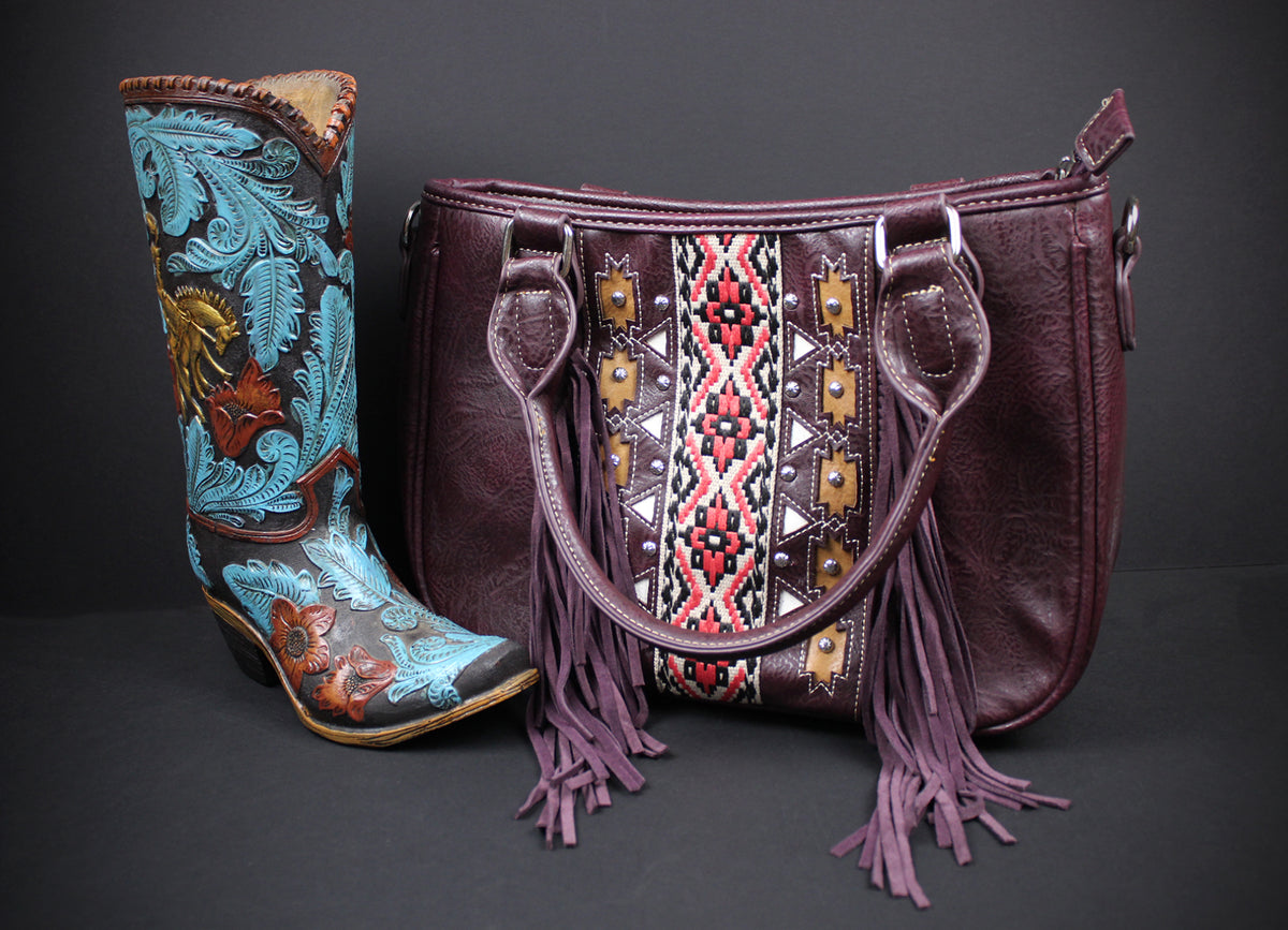 A dark burgundy leather handbag with purple fringe and a geometric embroidered panel, displayed next to a turquoise and brown embroidered cowboy boot against a dark gray background.