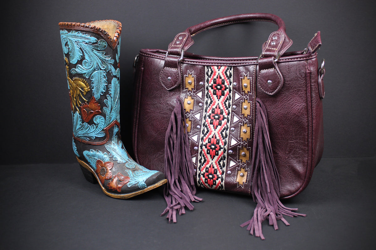 A western-style handbag in dark burgundy leather with purple fringe and a geometric embroidered panel, displayed next to a tall turquoise cowboy boot with floral tooling and gold-toned accents on a dark gray background.