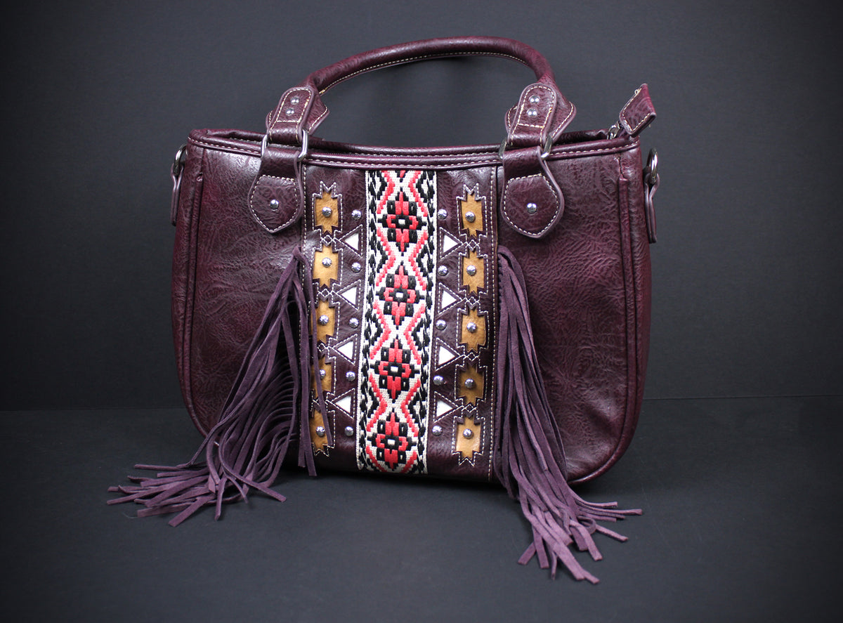 A dark purple faux leather handbag with a geometric patterned panel in red, white, and black, featuring purple fringe tassels hanging from the sides and top handles, set against a dark gray background.