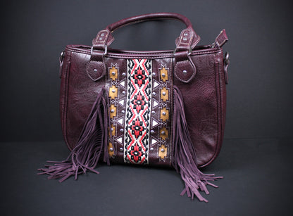A dark purple faux leather handbag with a geometric patterned panel in red, white, and black, featuring purple fringe tassels hanging from the sides and top handles, set against a dark gray background.