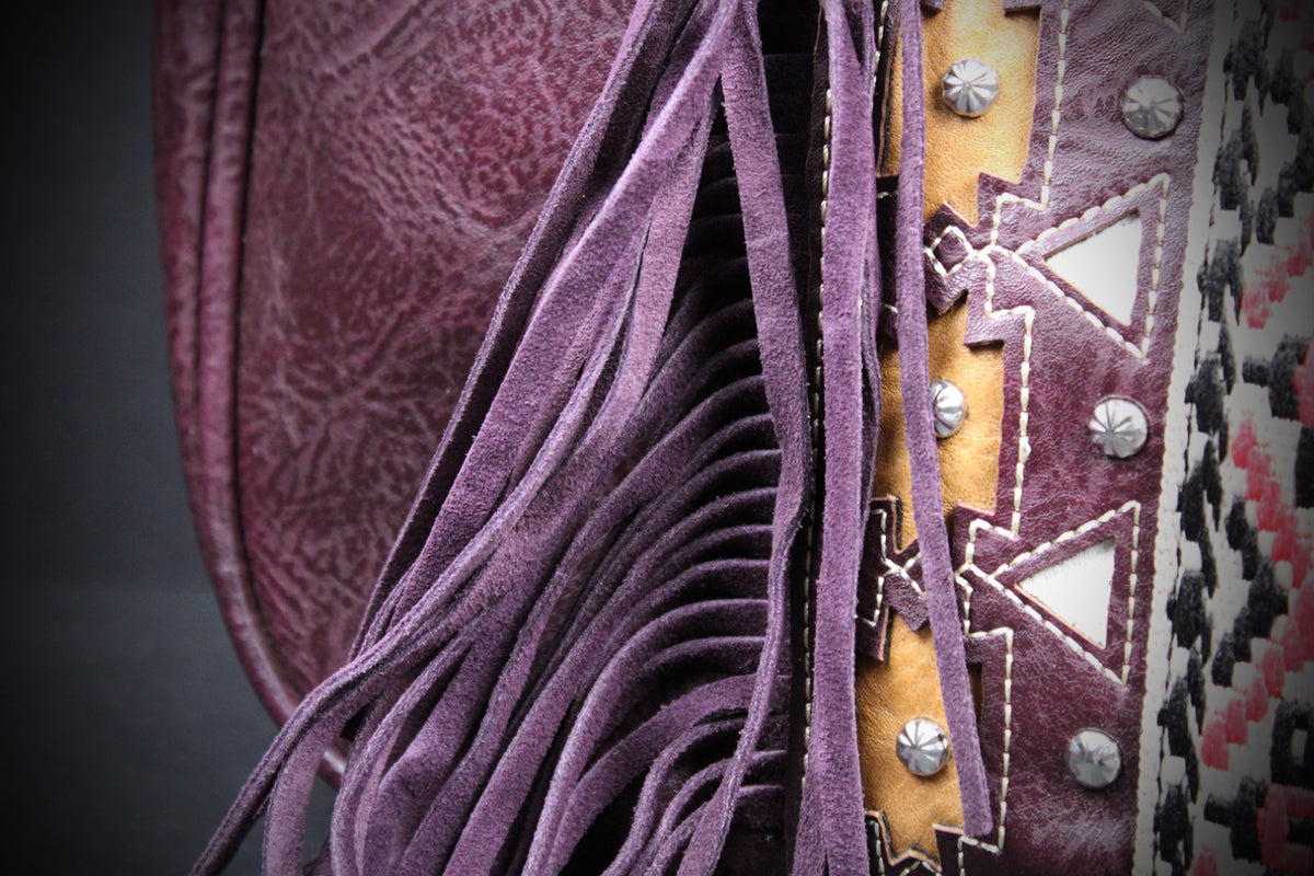 Close-up of a western-style leather fringe bag or holster in mauve-purple suede, showing layered fringe, a patterned leather panel with silver studs, and a woven textile detail against a dark background.