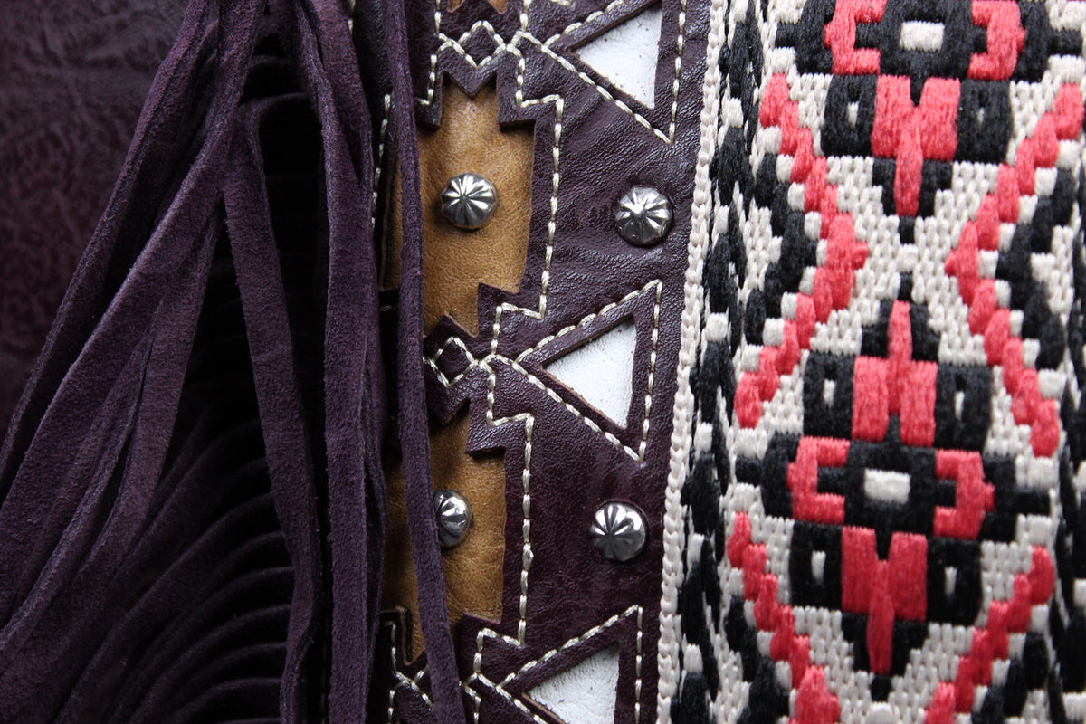 Close-up detail of a western-style textile accessory showing dark purple fringe, brown leather with white stitching and silver studs, and a black, red, and white geometric woven pattern.