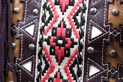 Close-up of a western-style textile or bag panel with a geometric embroidered pattern in red, black, and off-white, bordered by dark brown leather and tan leather sections with silver studs.