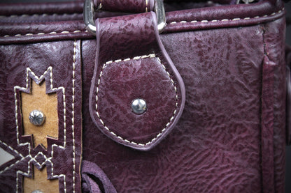 Close-up of a textured burgundy leather handbag featuring a snap-button strap with white contrast stitching and a tan geometric patch with silver studs.