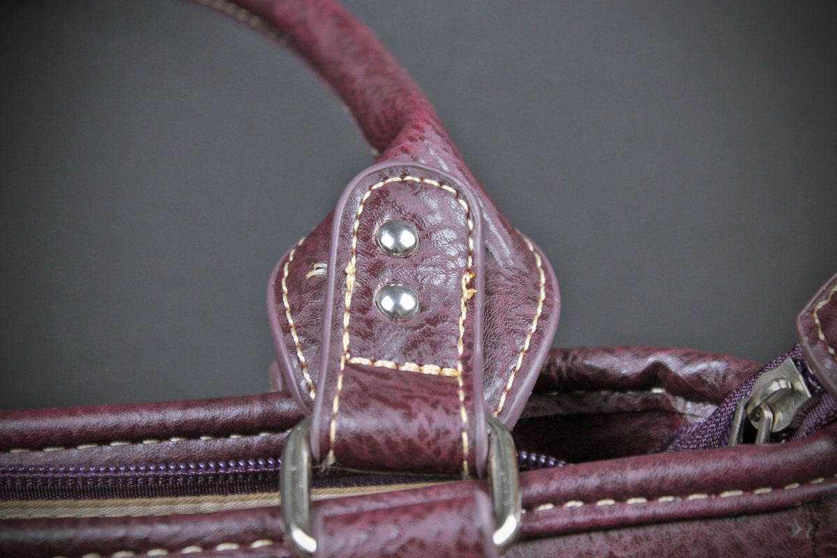 Close-up detail of a distressed burgundy leather handbag handle attachment with silver-tone rivets and metal hardware against a dark gray background.