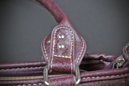 Close-up detail of a distressed burgundy leather handbag handle attachment with silver-tone rivets and metal hardware against a dark gray background.