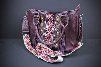 A burgundy faux leather handbag with fringe details and a wide, patterned woven shoulder strap featuring geometric red, black, white, and yellow designs, set against a dark gray background.