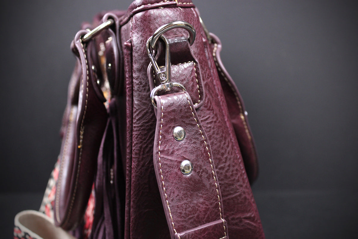 Close-up of a burgundy pebbled leather handbag showing the side profile, silver-tone hardware, and a patterned fabric lining or strap detail against a dark background.