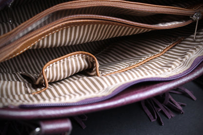 Close-up of the interior of a purple leather bag with a beige and brown striped fabric lining, showing zippers, a pocket, and fringe details.