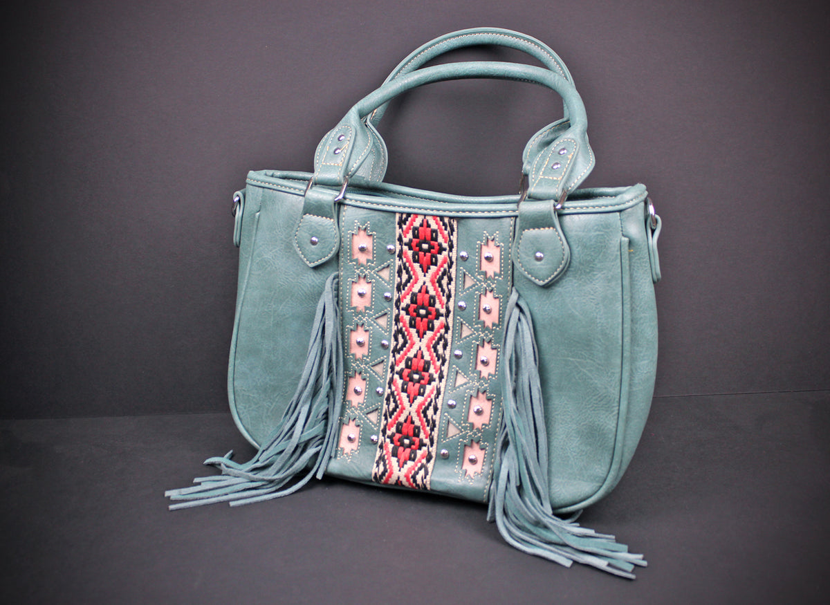 A sage green faux leather handbag with a central embroidered panel in red, pink, and white geometric patterns, featuring long fringes hanging from the bottom and dual top handles, set against a dark gray background.
