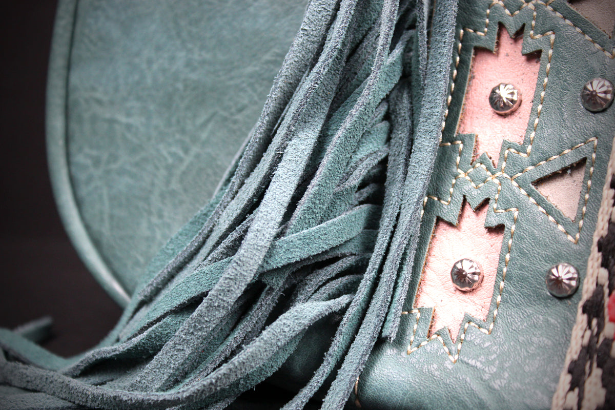 Close-up of a fringed leather accessory in a muted teal-green color, featuring layered suede-like fringe, a decorative stitched panel, and metal studs.