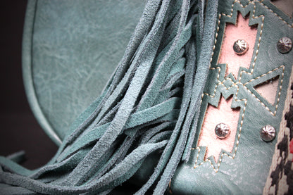 Close-up of a fringed leather accessory in a muted teal-green color, featuring layered suede-like fringe, a decorative stitched panel, and metal studs.