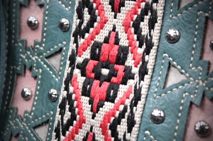 Close-up of a western-style garment or accessory featuring a central geometric embroidered panel in red, black, and off-white, bordered by teal leather trim with silver studs and a pink fabric backing.