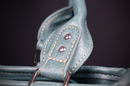 Close-up of a teal leather bag strap with contrast tan stitching and two metal rivets, set against a dark background.