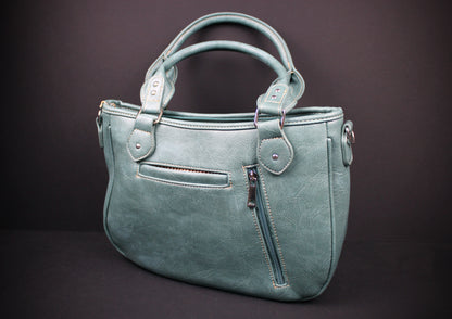 A dusty teal faux-leather handbag with dual top handles, a front zippered pocket, and a vertical zippered pocket on the front, set against a dark brown background.