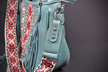 Close-up of a teal leather shoulder bag with fringe, silver studs, and a wide woven patterned strap in red, black, and cream, shown against a dark gray background.