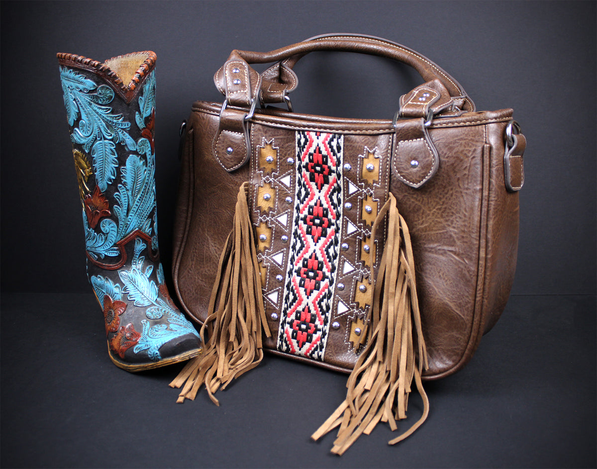 A brown leather handbag with tan fringe and a central embroidered geometric panel, displayed next to a tall embroidered cowboy boot on a dark studio background.