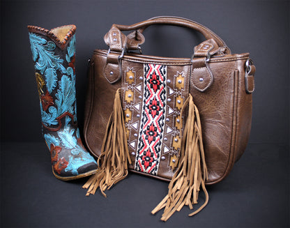 A brown leather handbag with tan fringe and a central embroidered geometric panel, displayed next to a tall embroidered cowboy boot on a dark studio background.