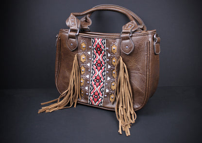 A brown faux-leather handbag with a western-style design, featuring a central embroidered panel with geometric patterns in red, black, and white, tan fringe tassels hanging from the sides, and a top handle, shown against a dark gray background.