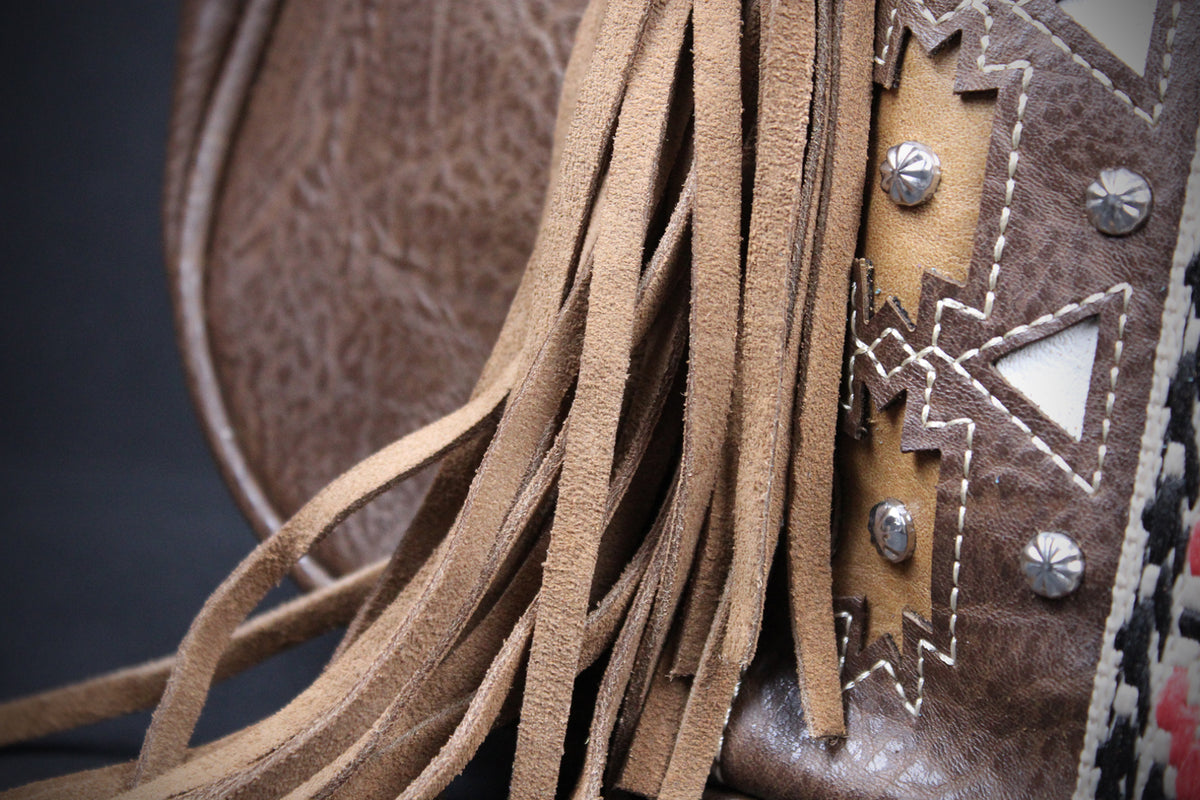 Close-up detail of a brown leather fringe bag with tan suede fringe, silver studs, and geometric white stitching.