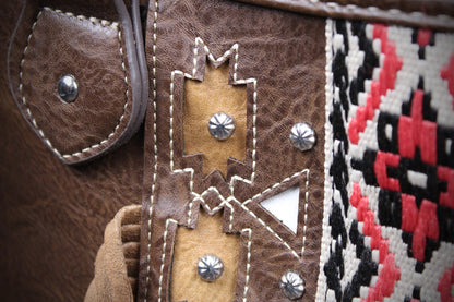 Close-up detail of a western-style bag showing brown leather with whipstitching, tan leather applique with silver studs, and a woven textile panel in black, white, and red geometric pattern.