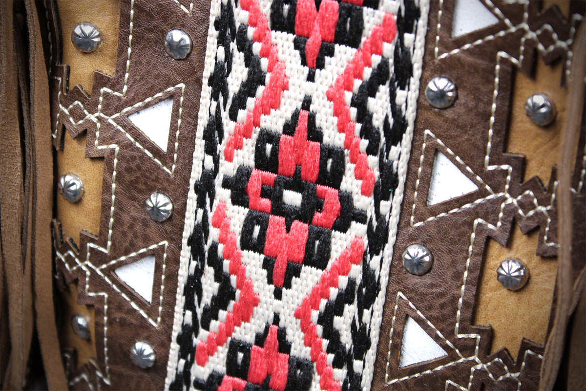 Close-up of a western-style textile or vest detail with brown suede-like panels, silver studs, and a central woven geometric pattern in red, black, and off-white.