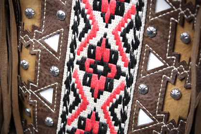Close-up of a western-style textile or vest detail with brown suede-like panels, silver studs, and a central woven geometric pattern in red, black, and off-white.