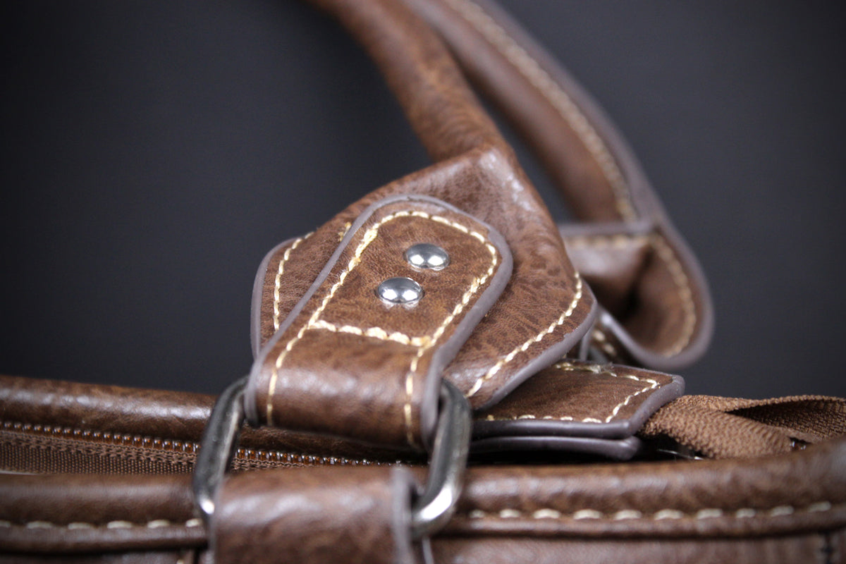 Close-up detail of a brown leather handbag strap with contrast stitching, metal rivets, and a silver-tone ring hardware against a dark background.