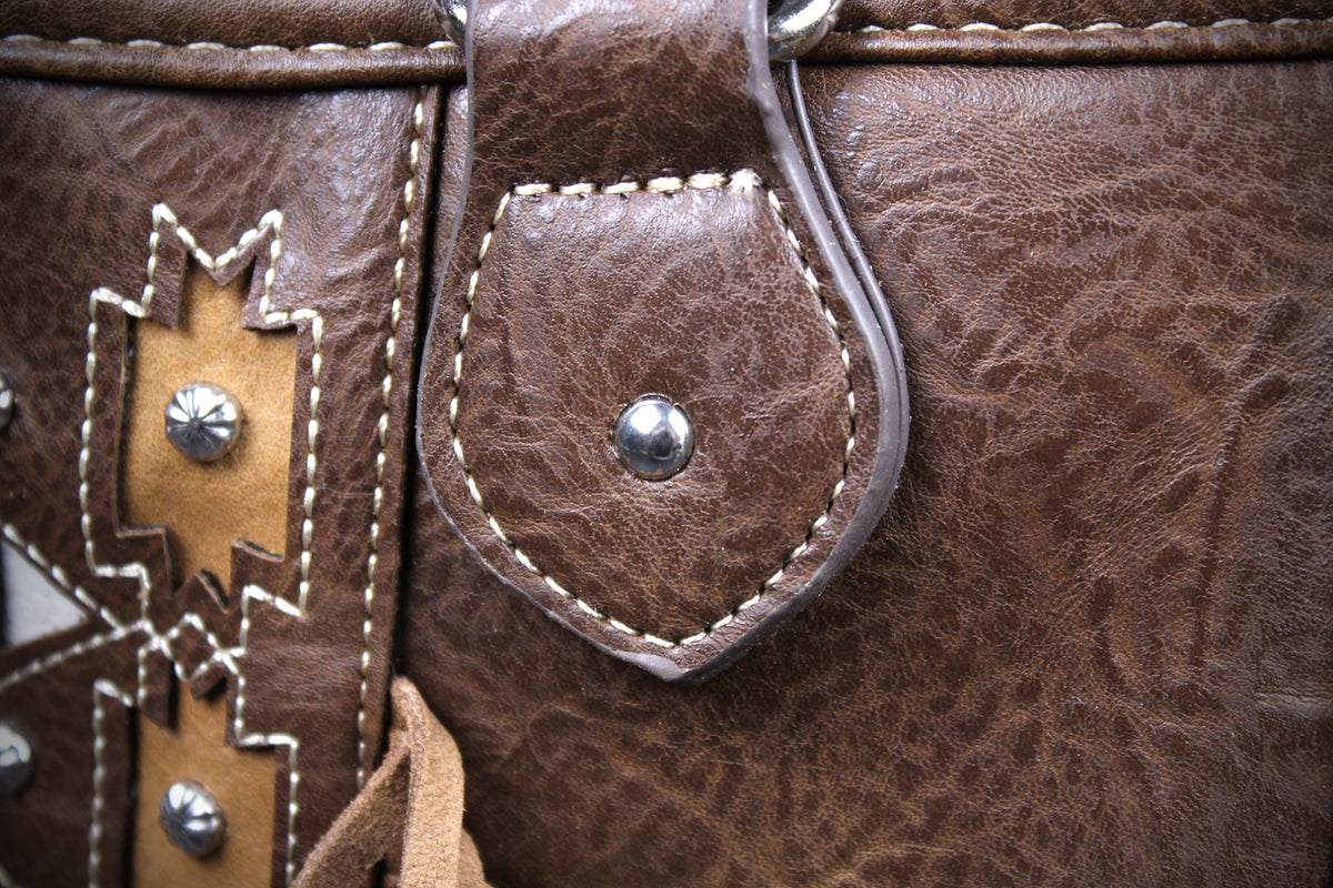 Close-up of a brown leather bag or holster showing textured leather panels, contrast stitching, a tan western-style stitched inlay with silver studs, and a small leather tab secured by a metal rivet.