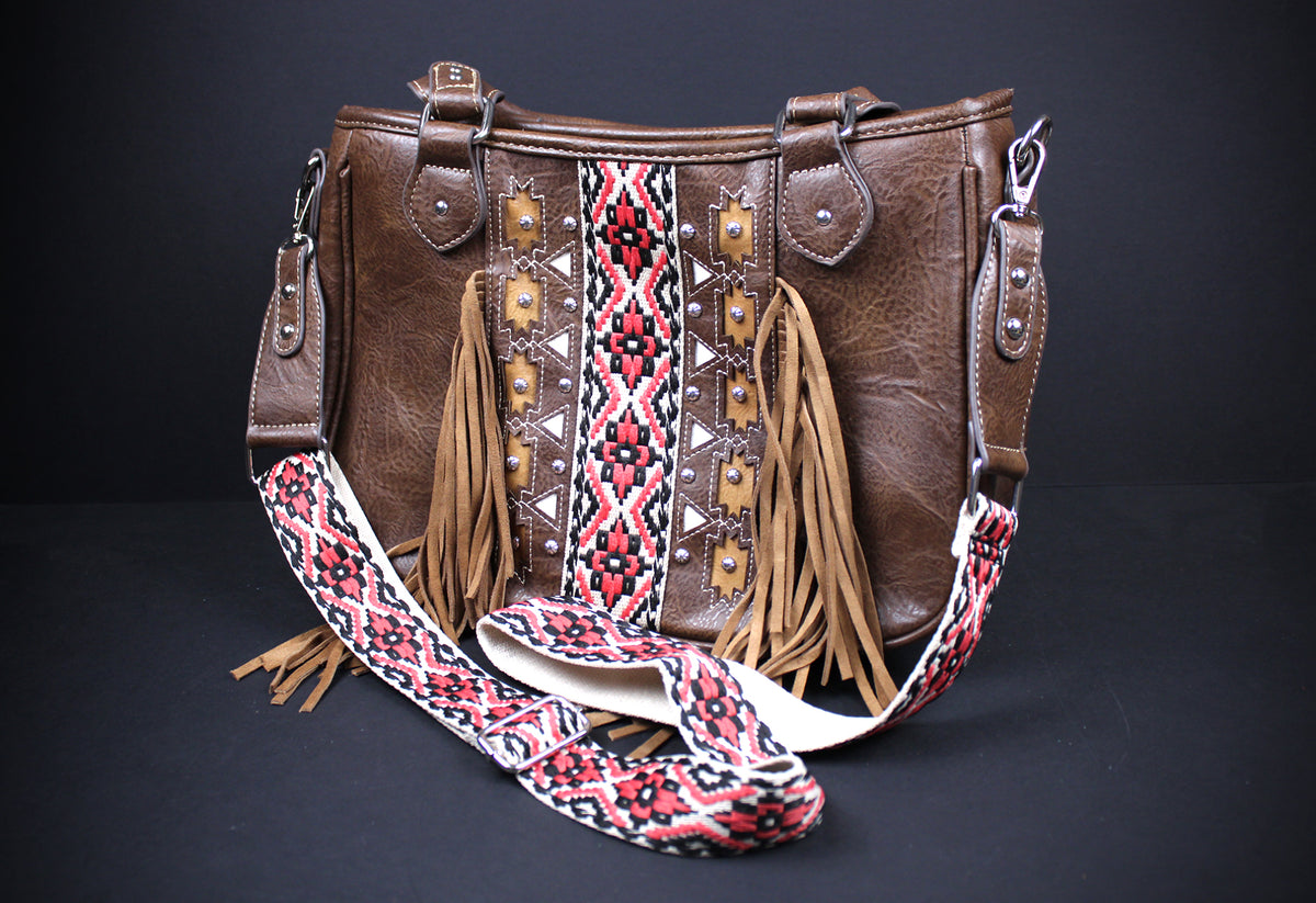 A brown faux-leather shoulder bag with a wide, patterned woven strap, long tan fringe, and geometric embroidered details on the front, set against a dark background.