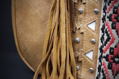 Close-up detail of a tan leather fringe accessory with geometric stitched panels and silver studs, next to a black, red, and white patterned textile.