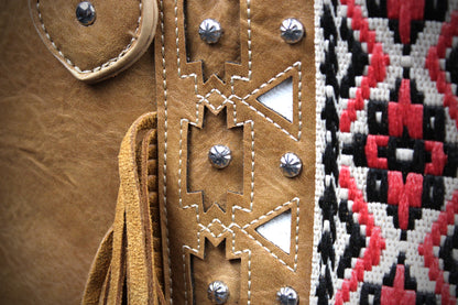 Close-up detail of a tan leather fringed garment or accessory, featuring geometric cutout stitching with silver studs and a black, red, and white woven textile pattern along the side.