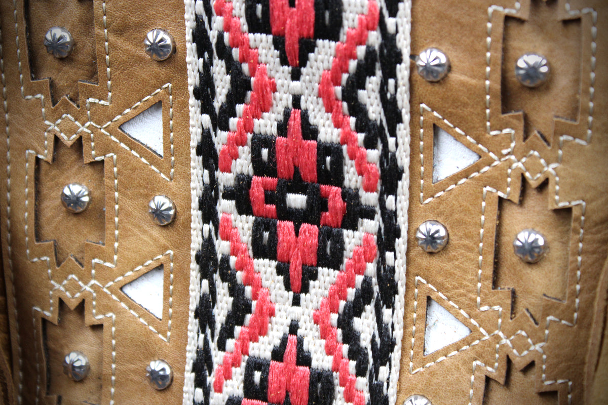 Close-up of a handcrafted leather item featuring a central woven geometric pattern in red, black, and white, flanked by tan leather panels with cutouts and silver-toned studs.