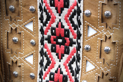 Close-up of a handcrafted leather item featuring a central woven geometric pattern in red, black, and white, flanked by tan leather panels with cutouts and silver-toned studs.