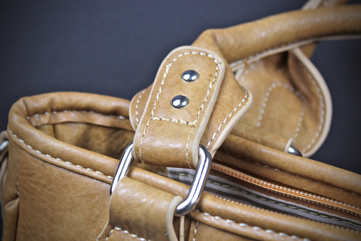 Close-up detail of a tan leather handbag showing the strap attachment with silver metal rings and contrast stitching against a dark background.