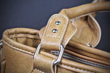 Close-up detail of a tan leather handbag showing the strap attachment with silver metal rings and contrast stitching against a dark background.